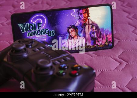 Dans cette illustration photo, le logo Wolf Among US 2 est affiché sur l'écran d'un smartphone, à côté d'une manette de jeu Banque D'Images