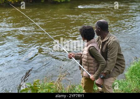 Vue arrière du père enseignant à son fils chaud à utiliser la canne à pêche alors qu'ils sont sur la rive des rivières et essaient d'attraper du poisson Banque D'Images