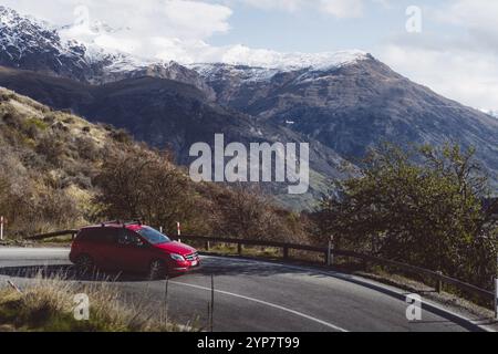 Une voiture rouge conduisant sur une route de montagne sinueuse par beau temps, Queenstown, Nouvelle-Zélande, Océanie Banque D'Images