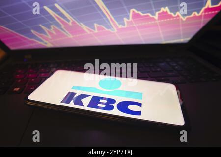 Dans cette illustration photo, le logo KBC Bank visible sur un écran de smartphone, avec une représentation graphique de la bourse en arrière-plan Banque D'Images