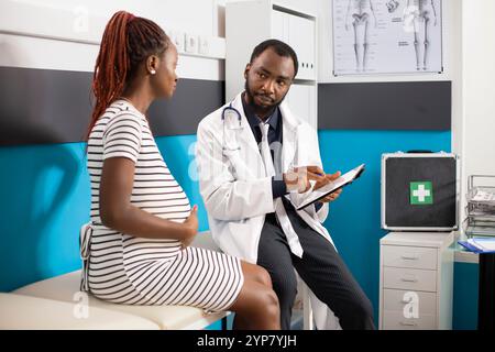 Médecin afro-américain est assis à côté de la dame enceinte tout en montrant les résultats médicaux sur l'appareil. Femme noire avec la main sur le ventre de grossesse écoute les conseils maternels du médecin tenant la tablette numérique Banque D'Images