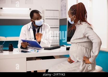 Médecin afro-américain et femme enceinte s'engagent dans la consultation prénatale dans le cabinet médical. Médecin parle de la santé maternelle tandis que la future mère écoute avec les mains reposant sur le ventre de la grossesse. Banque D'Images