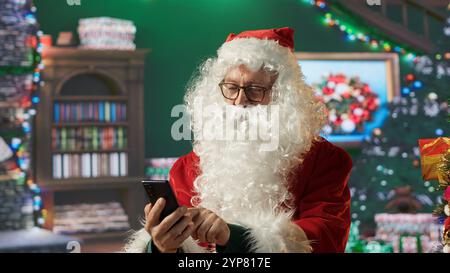 Saint Nick navigue sur le Web sur son téléphone portable, les listes de vérification et les emplacements pour la livraison sur la carte du monde. Cabine Pôle Nord avec lumières de Noël et magie, préparations de vacances. Caméra A. Banque D'Images