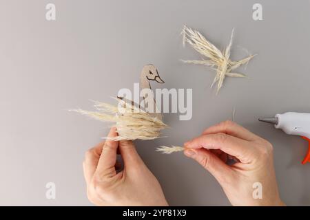 Mains fabriquant figurine de cygne à partir de carton et d'herbe de pampa à l'aide d'un pistolet à colle chaude sur un fond gris. Concept de projet de décoration DIY. passe-temps créatif c Banque D'Images