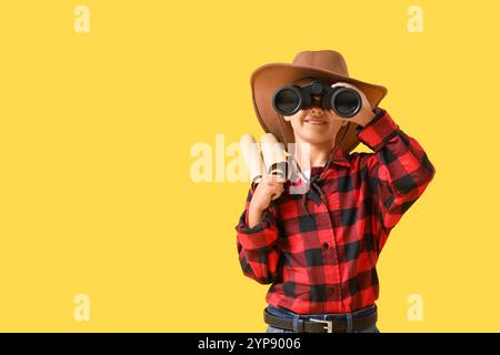 Mignon petit cow-boy avec carte au trésor regardant à travers des jumelles sur fond jaune Banque D'Images