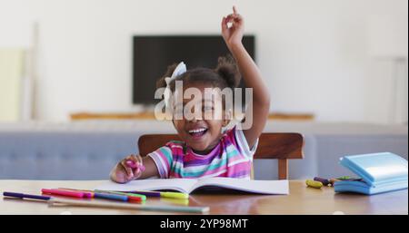 Heureuse fille afro-américaine levant la main pendant l'école en ligne à la maison Banque D'Images