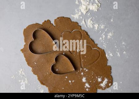 La pâte à biscuits est déroulée avec des couteaux en forme de cœur prêts à être cuits dans une cuisine chaude, parfaite pour les festivités hivernales. Banque D'Images