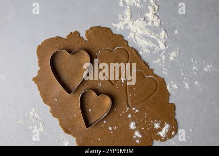 La pâte à biscuits est déroulée avec des couteaux en forme de cœur prêts à être cuits dans une cuisine chaude, parfaite pour les festivités hivernales. Banque D'Images