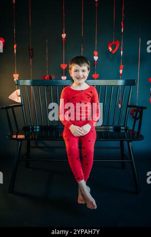 Garçon souriant en pyjama coeur rouge assis sur un banc noir Banque D'Images