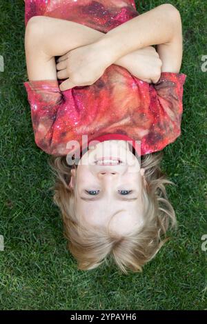 Sourire mignon garçon de 7 ans dans un T-shirt rouge se trouve sur l'herbe verte et regarde la caméra. Concept d'émotions positives, enfance heureuse. Haut vi Banque D'Images