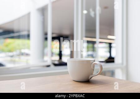 tasse à café blanche placée sur une table en bois près d'une fenêtre lumineuse, capturant un moment serein de simplicité moderne et de lumière naturelle. Parfait pour les vacances pr Banque D'Images