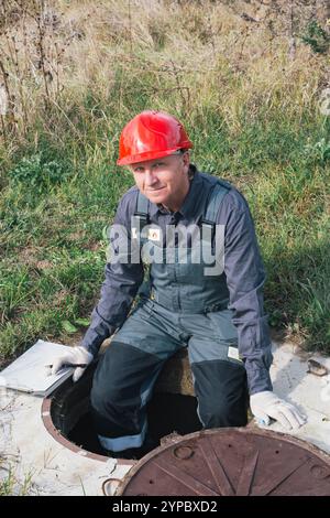 Travailleur dans le casque souriant tout en étant assis sur le puits d'eau libre. Entretien des puits, compteurs et tuyaux d'eau. Banque D'Images