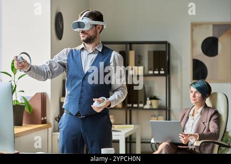 Jeune homme d'affaires moderne avec casque VR à l'aide de contrôleurs tout en faisant la présentation du nouveau projet contre collègue Banque D'Images