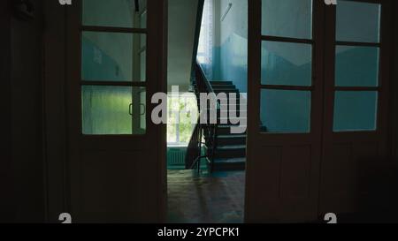 La lumière qui circule à travers des portes ouvertes illumine un couloir ombragé et un escalier dans une école vide le dernier jour Banque D'Images