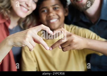 Enfant biracial jouant avec deux adultes et faisant des gestes en forme de coeur avec leurs mains, souriant et profitant du temps ensemble. Les aînés et les enfants embrassent des moments joyeux dans cette photo en gros plan Banque D'Images