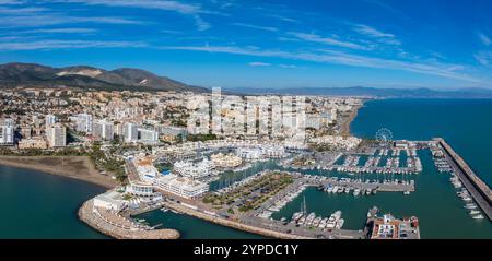 Vue aérienne de Benalmadena, Espagne, avec un port de plaisance animé avec des yachts, des plages de sable, une grande roue et des bâtiments blancs contre des collines ondulantes. Banque D'Images