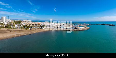 Vue aérienne de Benalmadena, Espagne, avec une plage de sable, un port de plaisance avec des bateaux, des bâtiments blancs et une grande roue contre un ciel clair et des montagnes. Banque D'Images