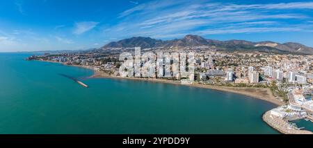 Vue aérienne de Benalmadena, Espagne, avec des bâtiments blancs et pastel, plage de sable, jetée, mer turquoise et collines luxuriantes sous un ciel bleu clair. Banque D'Images