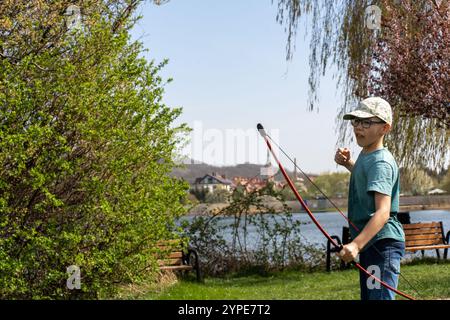 Jeune garçon pratiquant le tir à l'arc avec arc rouge près du lac pittoresque sur la journée ensoleillée, entouré d'une verdure vibrante et village pittoresque en arrière-plan. CON Banque D'Images