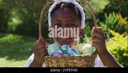 Fille afro-américaine souriante portant des oreilles de lapin de pâques tenant panier d'oeufs de pâques dans le jardin Banque D'Images