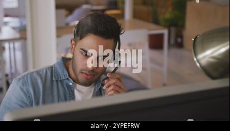Vue de face d'un homme biracial heureux travaillant dans un bureau créatif, portant un casque, parlant et souriant Banque D'Images