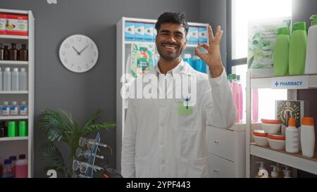 Beau jeune homme travaillant dans une pharmacie souriant et faisant des gestes bien à l'intérieur Banque D'Images