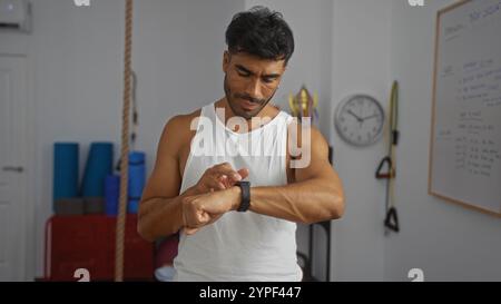 Jeune homme hispanique dans le gymnase regardant sa smartwatch, portant un débardeur blanc, avec un équipement d'exercice et une horloge en arrière-plan. Banque D'Images