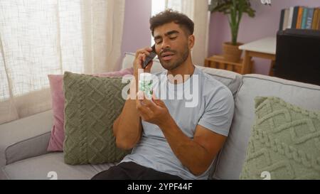 Homme hispanique assis dans un salon, inspectant la bouteille de médicament pendant un appel téléphonique, affichant un mélange équilibré de préoccupation et de concentration. Banque D'Images