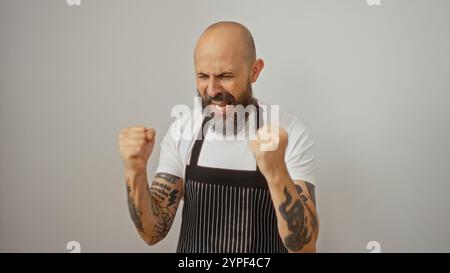 Bel homme hispanique avec une tête chauve et une barbe, portant un tablier, célébrant avec les poings serrés, isolé sur un fond blanc Banque D'Images
