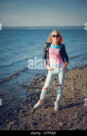 Jeune femme blonde portant des lunettes de soleil et une veste en cuir posant sur une plage de galets Banque D'Images