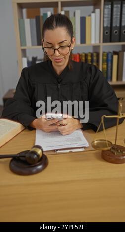 Jeune, hispanique, brune, femme juge dans une salle d'audience, portant des lunettes, utilisant un smartphone, entouré de livres de droit et de documents juridiques sur un de bois Banque D'Images