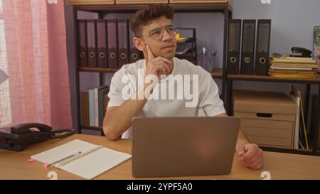 Beau jeune homme hispanique pensant tout en travaillant sur un ordinateur portable dans un bureau organisé orné de dossiers et d'étagères. Banque D'Images