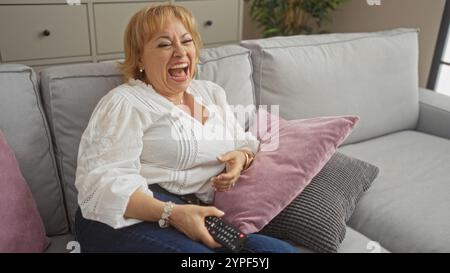 Une belle femme caucasienne âgée rit joyeusement tout en étant assise sur un canapé dans un salon confortable, tenant une télécommande dans sa main. Banque D'Images