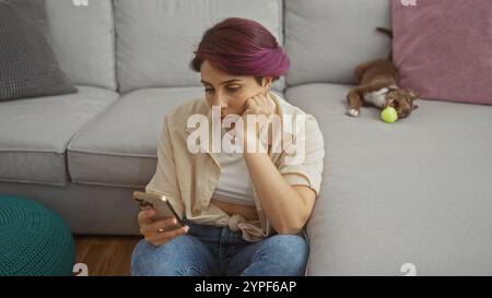 Jeune femme belle dans le salon d'un appartement, assise sur un canapé à l'aide d'un smartphone pendant que son chien joue avec une balle de tennis. Banque D'Images