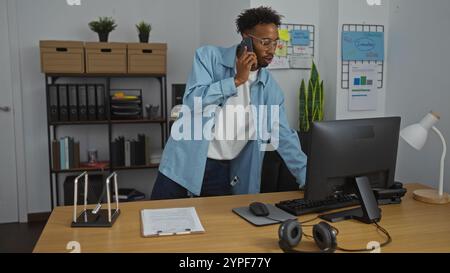Un jeune afro-américain avec une barbe parle au téléphone alors qu'il travaille à un ordinateur dans un bureau moderne. Banque D'Images