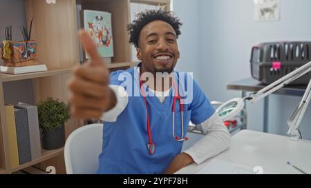 Beau jeune homme afro-américain avec une barbe portant des gommages et un stéthoscope donnant un pouce vers le haut dans un lieu de travail de clinique vétérinaire. Banque D'Images