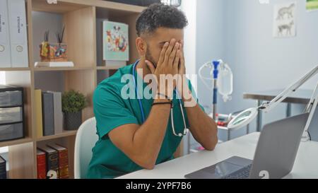 Portrait d'un jeune vétérinaire hispanique dans une clinique vétérinaire, paraissant stressé alors qu'il était assis à un bureau avec ses mains couvrant son visage. Banque D'Images