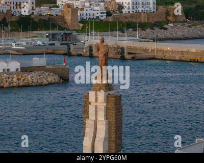 Une statue se dresse sur un piédestal de pierre dans le port de Tarifa, en Espagne. Les bâtiments blancs de la ville et les anciens murs de fortification sont visibles en arrière-plan. Banque D'Images