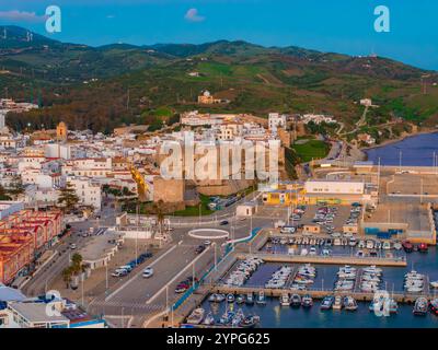 L'image capture une vue aérienne de Tarifa, en Espagne, mettant en évidence le château de Guzman, les bâtiments blanchis à la chaux, les collines verdoyantes, et un port animé avec bo Banque D'Images