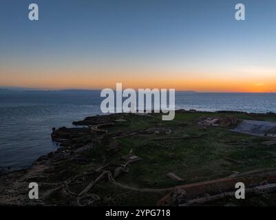 L'image montre une vue aérienne de la côte de Tarifa au coucher du soleil, avec un phare sur un éperon rocheux, des chemins sinueux, des ruines dispersées, et un vaste Banque D'Images