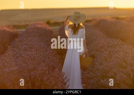 Lavender Field Woman Sunset : femme en robe blanche, chapeau de paille, explore un champ de lavande pittoresque pendant le coucher du soleil, appréciant la beauté sereine. Banque D'Images