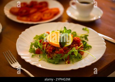 Tartare de thon décoré avec roquette, demi-tranches d'orange et de citron vert, et un brin de menthe repose sur une assiette blanche, la table en bois et d'autres options de petit déjeuner Banque D'Images
