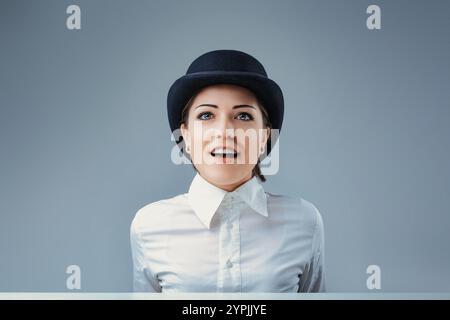 Portrait d'une jeune femme d'affaires surprise regardant vers le haut tout en portant un chapeau melon et une chemise blanche, isolée sur un fond gris, exprimant amazeme Banque D'Images