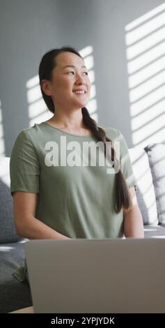 Plan vertical d'une femme asiatique utilisant un ordinateur portable et souriant par la fenêtre Banque D'Images