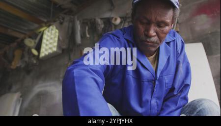 Vue de face d'un batteur à panneaux masculin senior africain dans un atelier du township, nettoyant un whee en alliage Banque D'Images