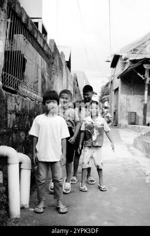 Bogor, 24 novembre 2024. Quatre jeunes enfants debout ensemble dans une ruelle étroite, souriant et regardant autour dans un cadre de quartier animé Banque D'Images