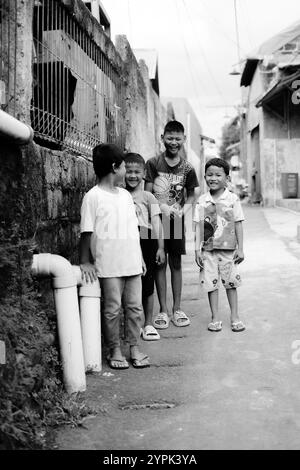 Bogor, 24 novembre 2024. Quatre jeunes enfants debout ensemble dans une ruelle étroite, souriant et regardant autour dans un cadre de quartier animé Banque D'Images