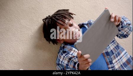 Heureux garçon afro-américain utilisant la tablette couchée sur le sol à la maison, ralenti Banque D'Images