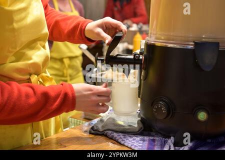 Dans un étal au marché de Noël, une jeune femme remplit du lait de poule chaud dans une tasse en verre givré, emploi saisonnier avec des boissons de vacances traditionnelles, selecte Banque D'Images