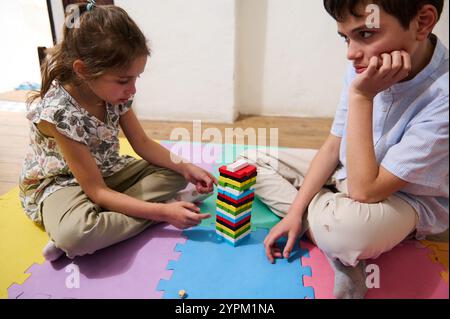 Deux enfants assis sur le sol concentrés sur un jeu d'empilement coloré. La fille place soigneusement un bloc pendant que le garçon regarde attentivement. C'est une scène de c. Banque D'Images